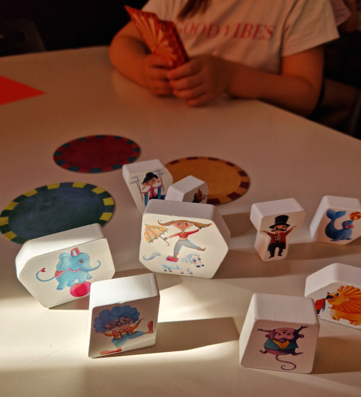 Białe elementy gry stojące na stoliku. W tle dziecko trzymające karty w ręku. 