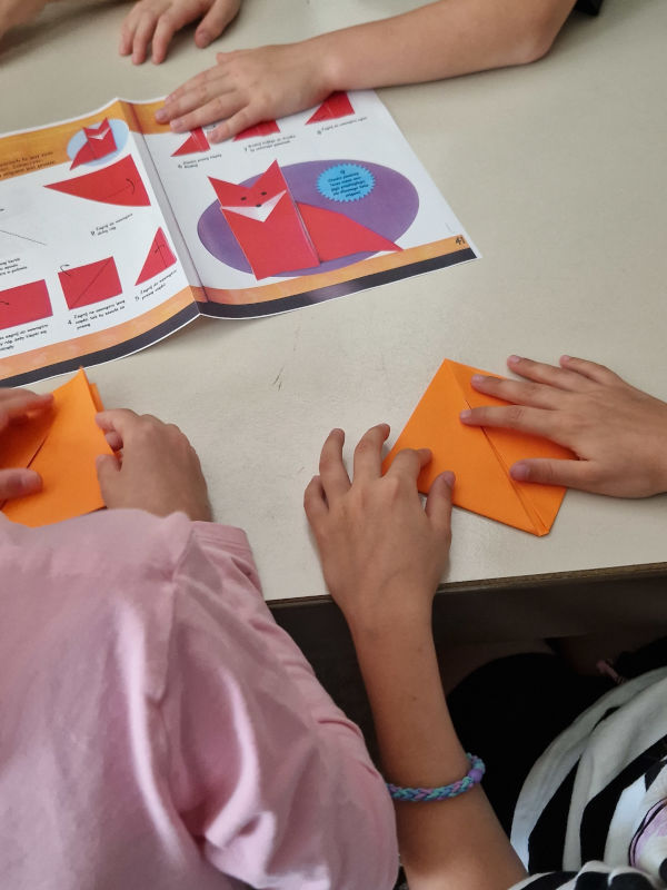 Ręce dzieci, które składają pomarańczowego lisa, wzorując się na wydrukowanym przykładzie. 