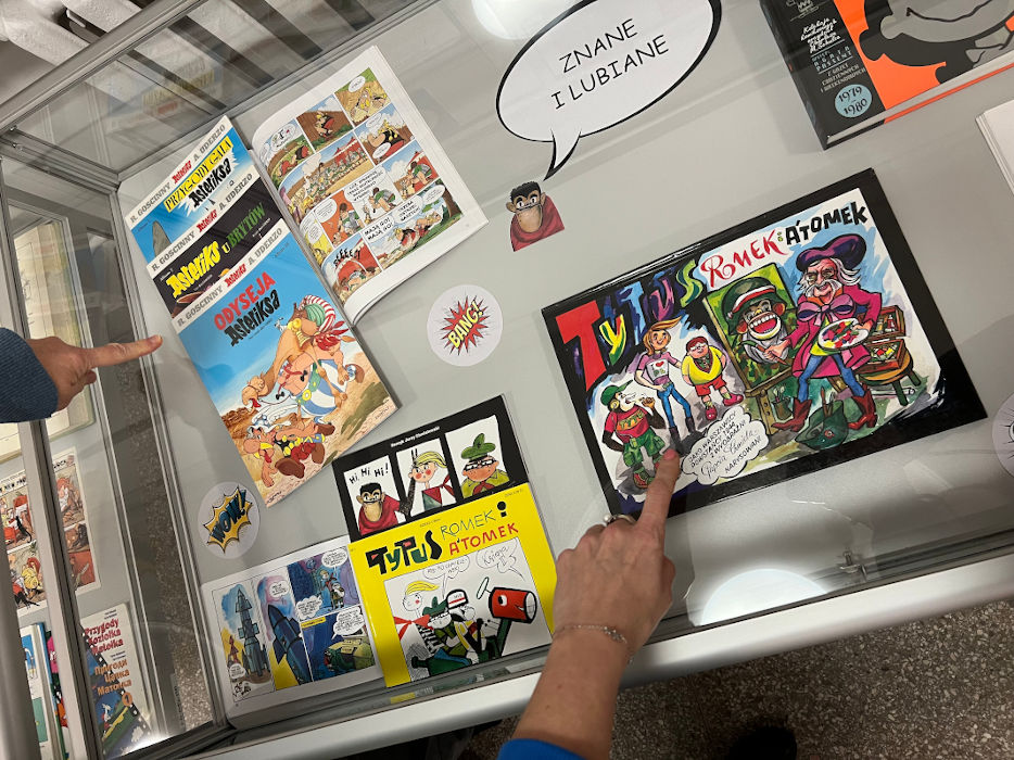 Dwie osoby wskazują palcami na komiksy w gablocie "Znane i lubiane"
