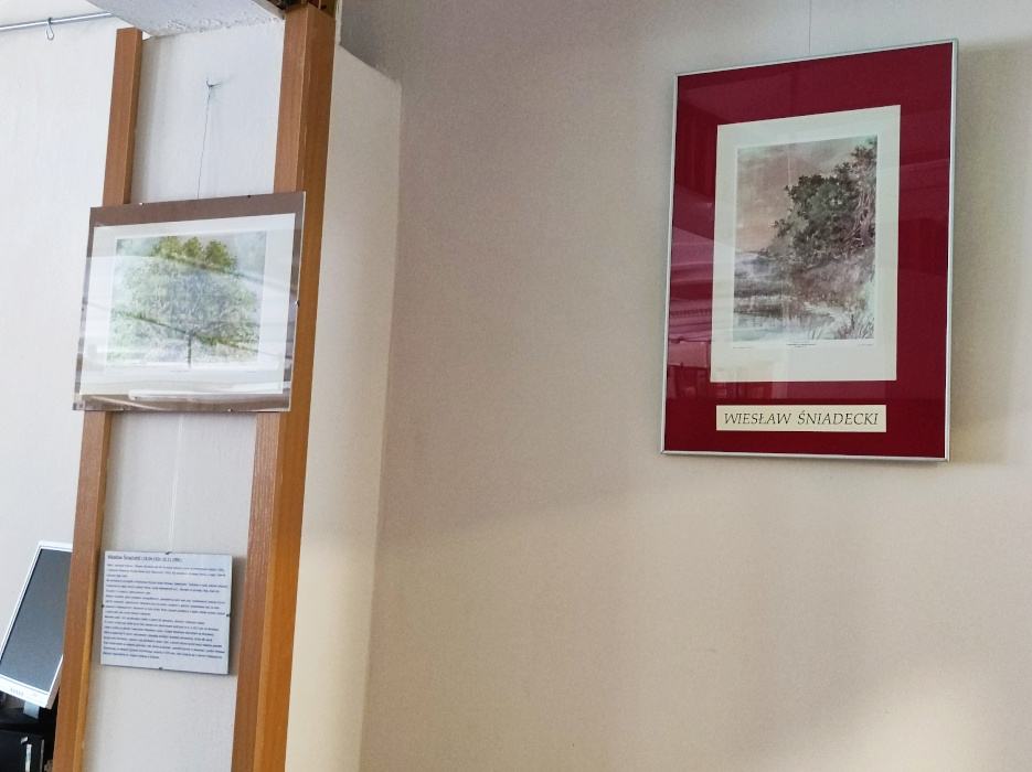 Dwa obrazy Wiesława Śniadeckiego wiszące na ścianie. Jeden na czerwonym tle przedstawiający drogę leśną zimą, drugi drzewo liściaste na jasnym tle