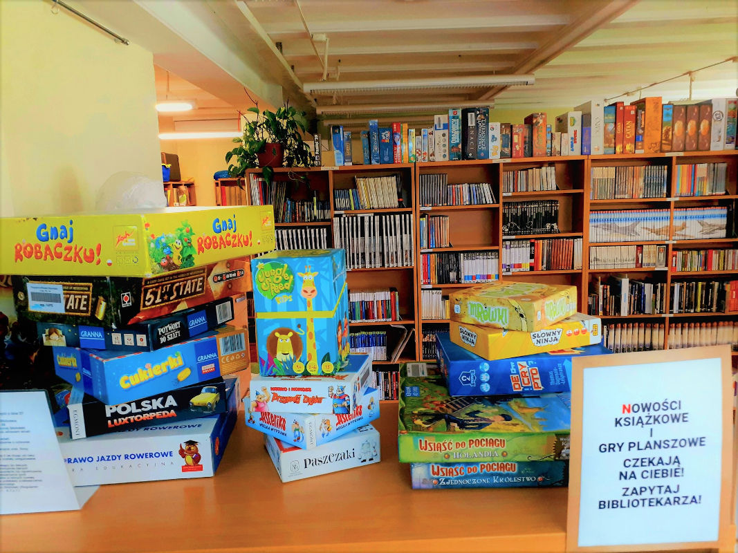 Kolorowe pudełka gier planszowych ułożone na sobie, w tle regały z książkami.